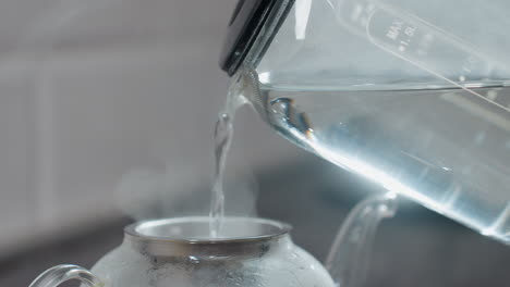 close up of person pouring hot water into glass kettle with metal filter, steam rising as water fills kettle, showing process of making hot tea or herbal infusion in kitchen setting