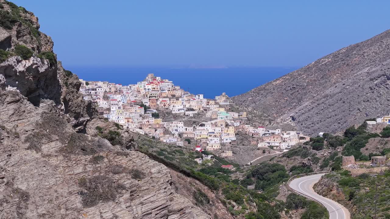 Aerial drone footage revealing the traditional Olympos village of Karpathos behind a hill in the foreground, showcasing the island’s stunning landscape and authentic Greek beauty