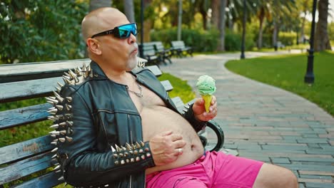 bald rocker in studded jacket and pink shorts savoring ice cream while sitting on park bench, embodying carefree summer leisure and alternative style