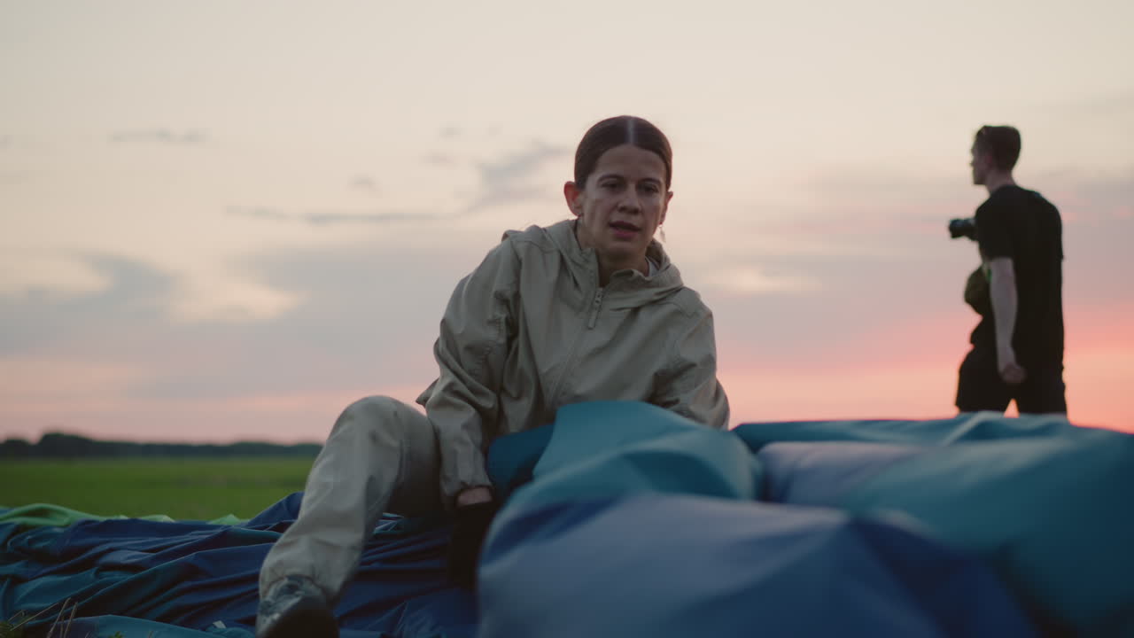 lady guiding deflated hot air balloon into parked position on grassy field at dusk after flight capturing teamwork careful handling of vibrant fabric against soft sunset horizon