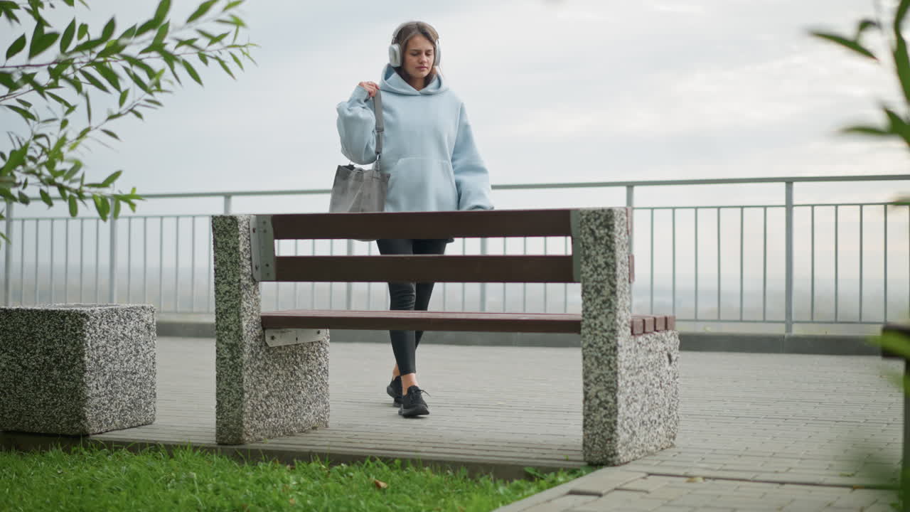 Lady walking toward concrete bench, carrying bag and wearing headphones, sitting carefully on bench in outdoor park, ideal for videos featuring relaxation, outdoor leisure, and tranquil moments