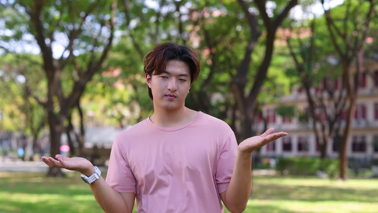 Một thanh niên châu Á mặc áo phông hồng nhún vai với hai tay mở, trông bối rối ngoài trời