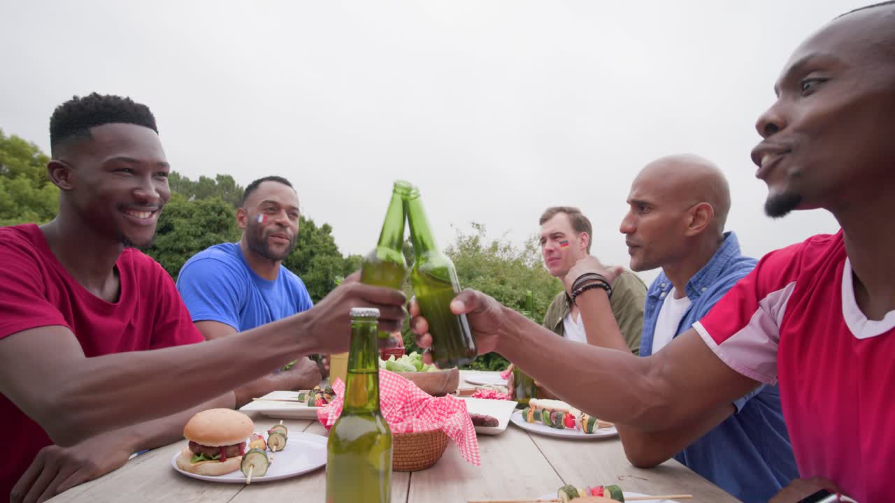 Amici uomini di diverse etnie appoggiati a un tavolo da picnic, un uomo prende la fotocamera per immortalare un brindisi con bottiglie verdi