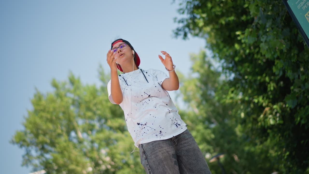 adolescente asiático en parque urbano ajustándose las gafas de sol, energía de creador de contenido con gorra informal y pose a la moda, toma en movimiento y expresiones juguetonas, vídeo vlog espontáneo, árboles de verano y fondo bokeh
