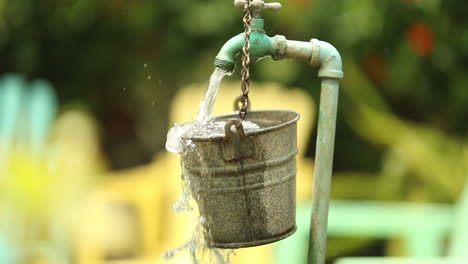 water from a garden spout pours and spills over a hanging bucket while the camera approaches with colored lounge chairs in the background