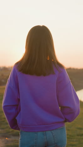 woman walks along riverbank at sunset. brunette female tourist enjoys quiet evening at spring riverside. lady in casual clothes at countryside