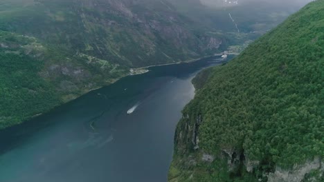 toma aérea en cámara lenta del fiordo de geiranger, noruega, con un barco en movimiento y una ciudad local en el fondo