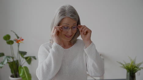 senior woman with gray hair and white shirt looking at camera smiling and putting on eyeglasses