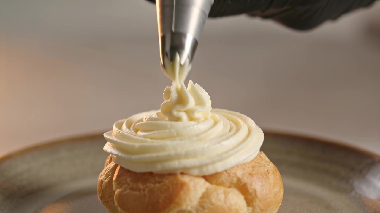 Piping gloved baker pressing piping bag at workstation, decorating cream puff with metal star tip