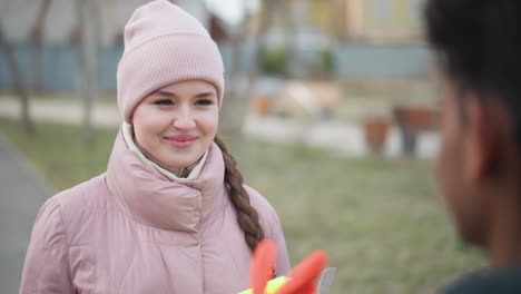 smiling woman in pink winter jacket and matching beanie with long braid walking outdoors in park on paved path towards person in foreground during early spring with grass and bare trees in background