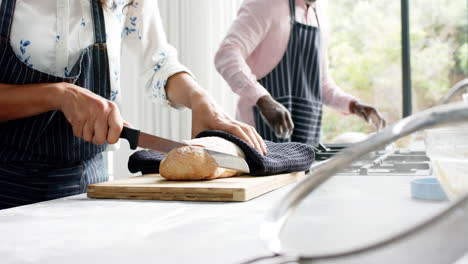 Mischlingspaar-Trägt-Schürzen-Und-Schneidet-Frisches-Brot-In-Der-Küche,-Zeitlupe