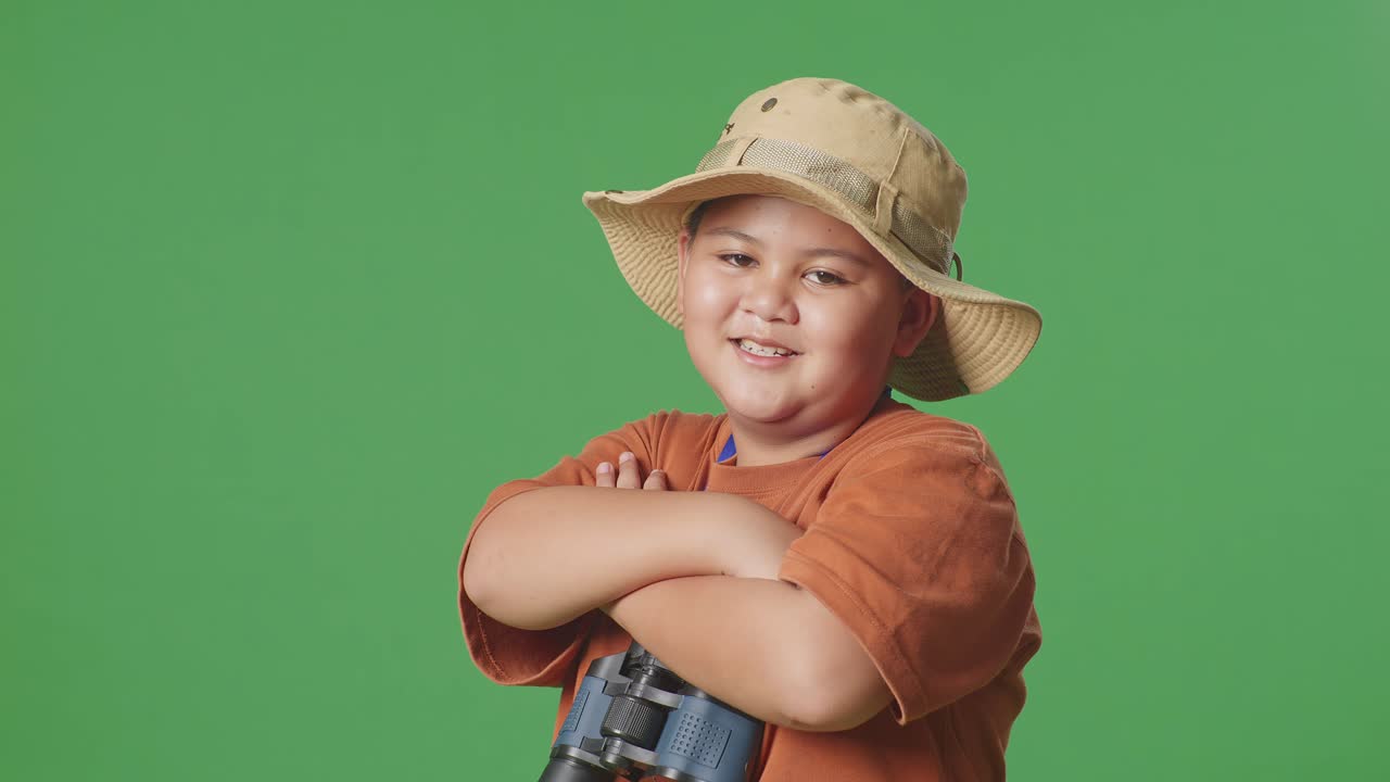 niño turista asiático con un sombrero y binoculares cruzando los brazos y sonriendo a la cámara mientras está de pie en el fondo de la pantalla verde. niño investigador examina algo, aventura de viaje, vista lateral de cerca
