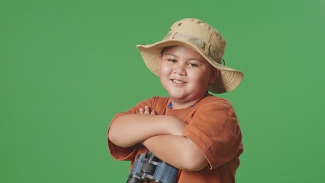 asian tourist boy with a hat and binoculars crossing arms and smiling to camera while standing on the green screen background. boy researcher examines something, travel adventure, close up side view