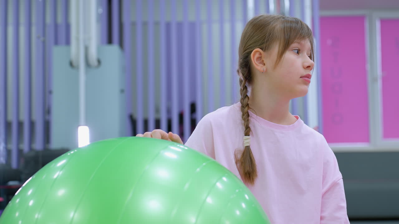 Pembe giyimli genç kız spor salonunda yeşil denge topunun yanında düşünceli bir şekilde oturuyor, arka planda mor ve pembe dekorlu, yumuşak zeminli ve egzersiz ekipmanlı modern bir fitness stüdyosu yer alıyor