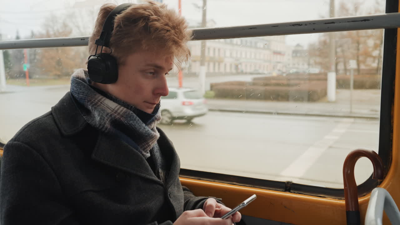 Jonge man luistert tijdens woon-werkverkeer. Jonge man bezig met mobiel apparaat tijdens stadsrit. Stedelijke jonge man met koptelefoon plant nauwgezet videocontent tijdens tramrit.