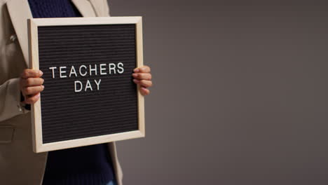 Close-Up-Studio-Shot-Of-Female-Teacher-Standing-Against-Grey-Background-Holding-Notice-Board-Reading-Teachers-Day-1