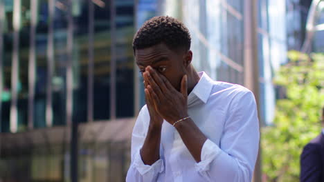 joven hombre de negocios cansado con mangas de camisa frotando los ojos de pie fuera de las oficinas en el distrito financiero de la ciudad de londres, reino unido