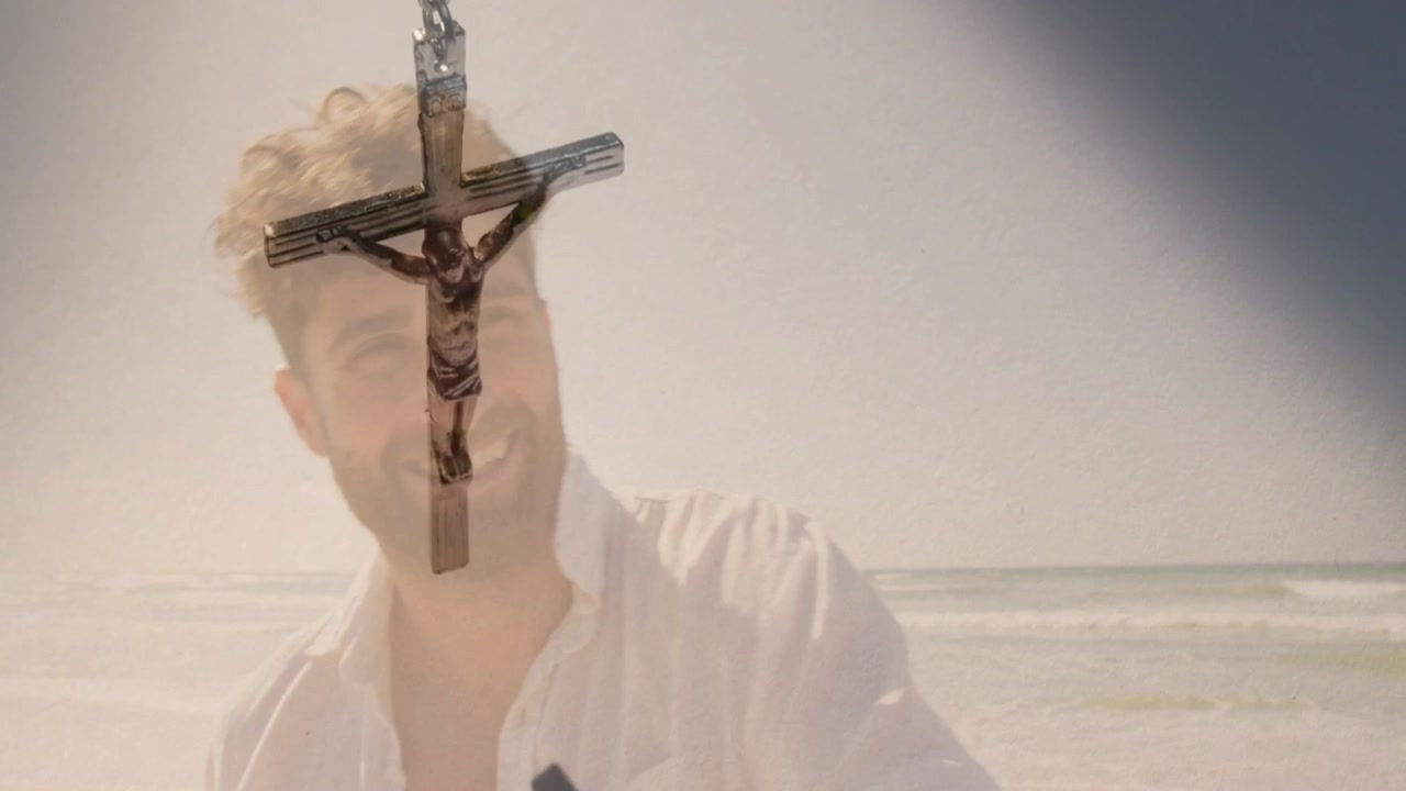 Man smiling behind crucifix pendant swinging from breeze, sunhat overlay fading, showcasing jewelry