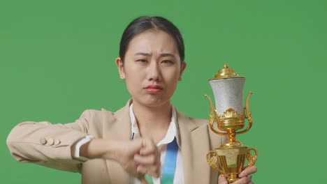 close up of asian business woman in a suit with a gold medal and trophy showing thumbs down gesture to ridicule the loser on green screen background in the studio