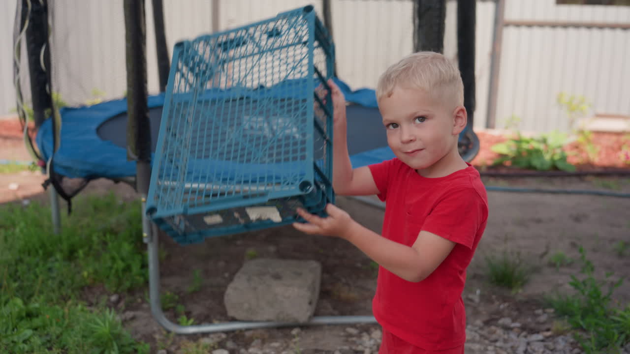 White Boy Inspecting Blue Plastic Crate Red Shirt Trampoline In Background Lifting And Turning Crate Curious Exploration Tactile Play Backyard Discovery Focused Expression Resourceful Hands Warm