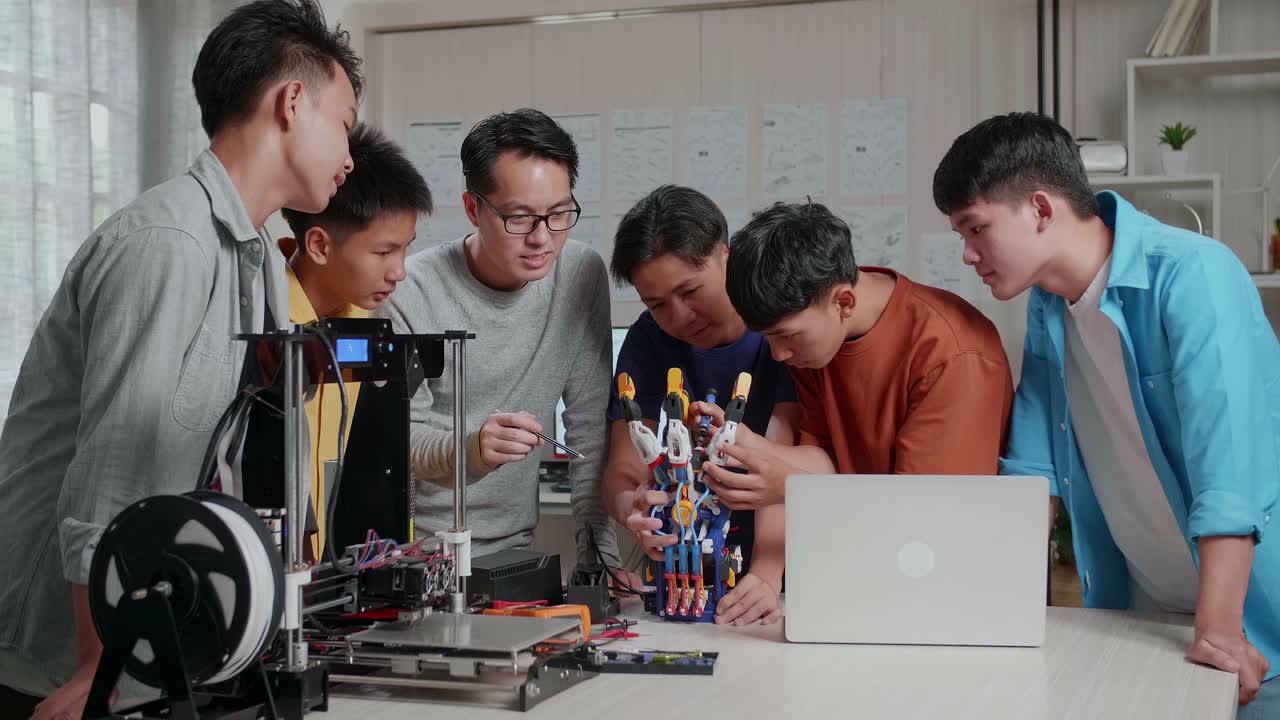 노트북과 3d 프린팅을 가진 아시아 남자가 학교에서 사이보그 손을 수리하는 소년들을 가르치고 있습니다.