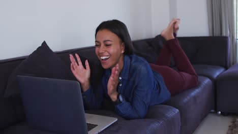 mujer de raza mixta acostada en el sofá usando computadora portátil y riendo