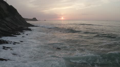 Drone-flying-at-low-altitude-over-ocean-waves-breaking-on-rocks-with-sunset-in-background,-Pyrenees-Atlantiques-in-France