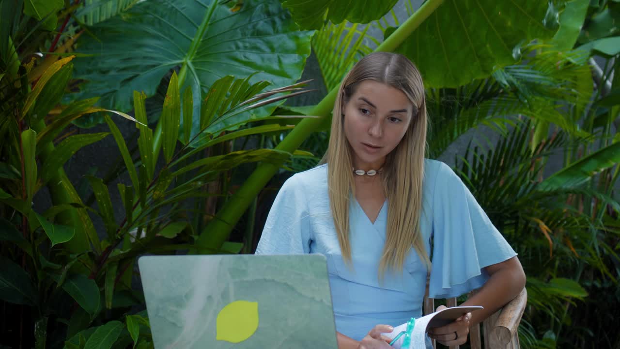 una chica en un vestido azul remotamente en línea trabajando detrás de una computadora portátil y mirando a la pantalla el patio trasero con plantas verdes