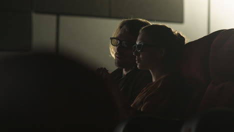 vista lateral de una pareja con anteojos 3d sentados en el cine mientras ven una película y comen palomitas de maíz con las luces apagadas