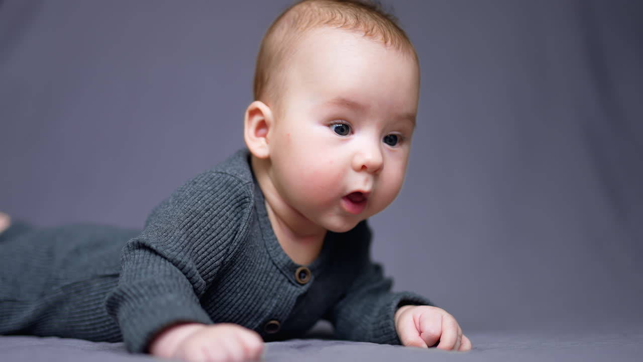 Nice sweet baby boy in grey clothes lies on belly. Cute toddler looks sideways and opens little mouth. Close up. Grey backdrop.