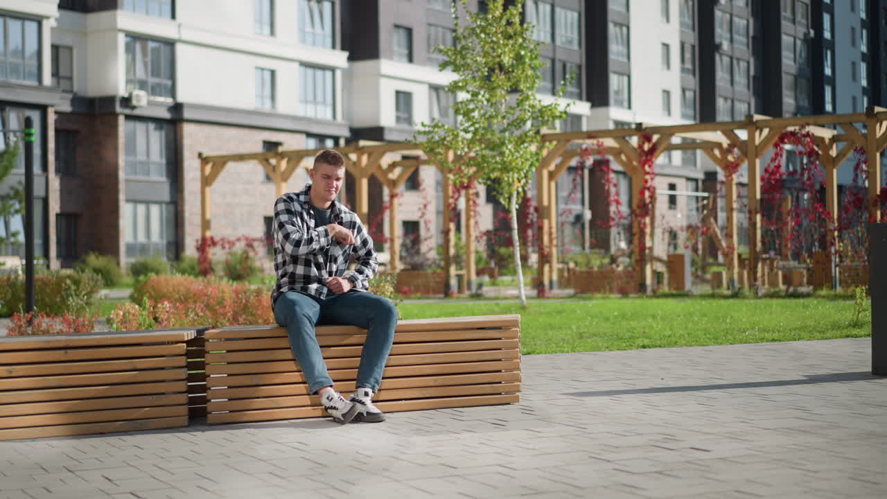young man dressed in checkered shirt sits on wooden bench in sunny outdoor plaza beside modern office complex pulling item from pocket while plants sway gently in breeze across landscaped area