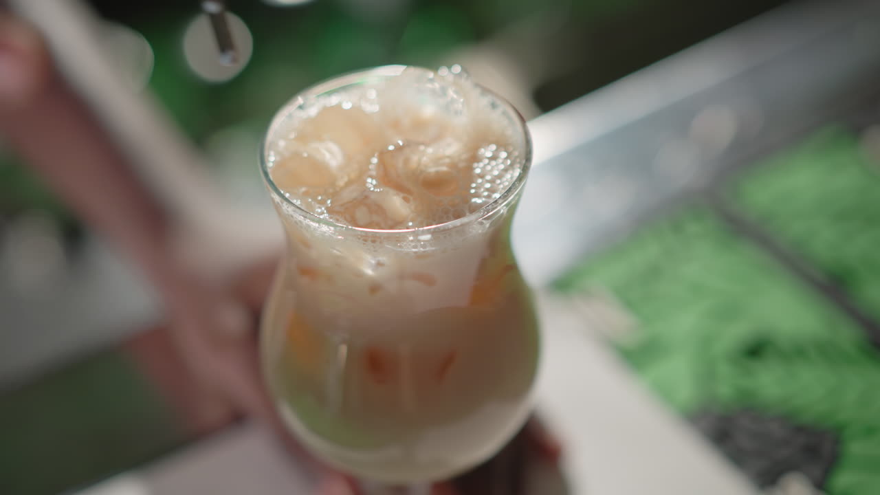 Close-up of creamy iced drink with bubbles rising as bartender finishes pouring from shaker. Ice cubes float, creating a refreshing, frothy look with vibrant bar background