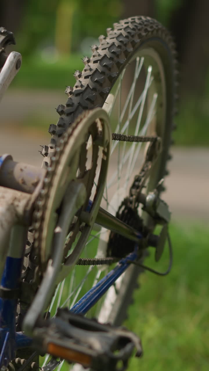 vista parcial de cerca de la mano de alguien girando el pedal de la bicicleta a la izquierda, centrándose en la cadena y el sistema de engranajes, el fondo suavemente borroso incluye una vista de la vegetación y el edificio