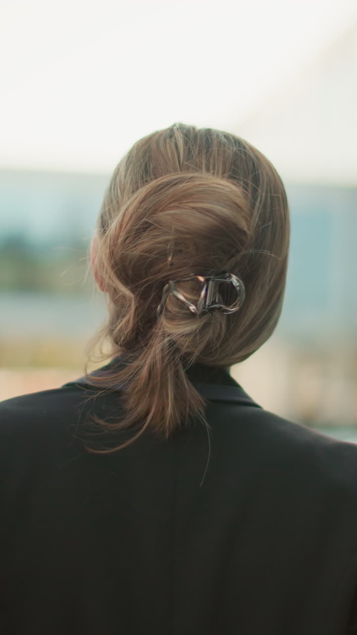 Back view of lady walking with red folder and coffee cup through city parking lot with blurred background of parked cars and glass office building on bright day