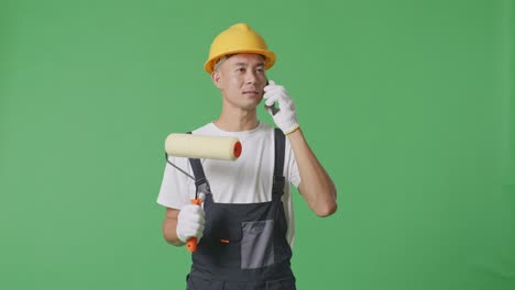 Asian Man Painter Wearing Safety Helmet Talking On Smartphone And Looking Around While Standing In The Green Screen Background Studio