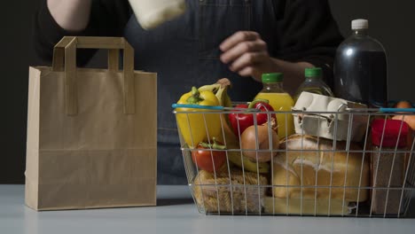 studio shot of shop worker packing basic food items in supermarket wire shopping basket into paper bag 1