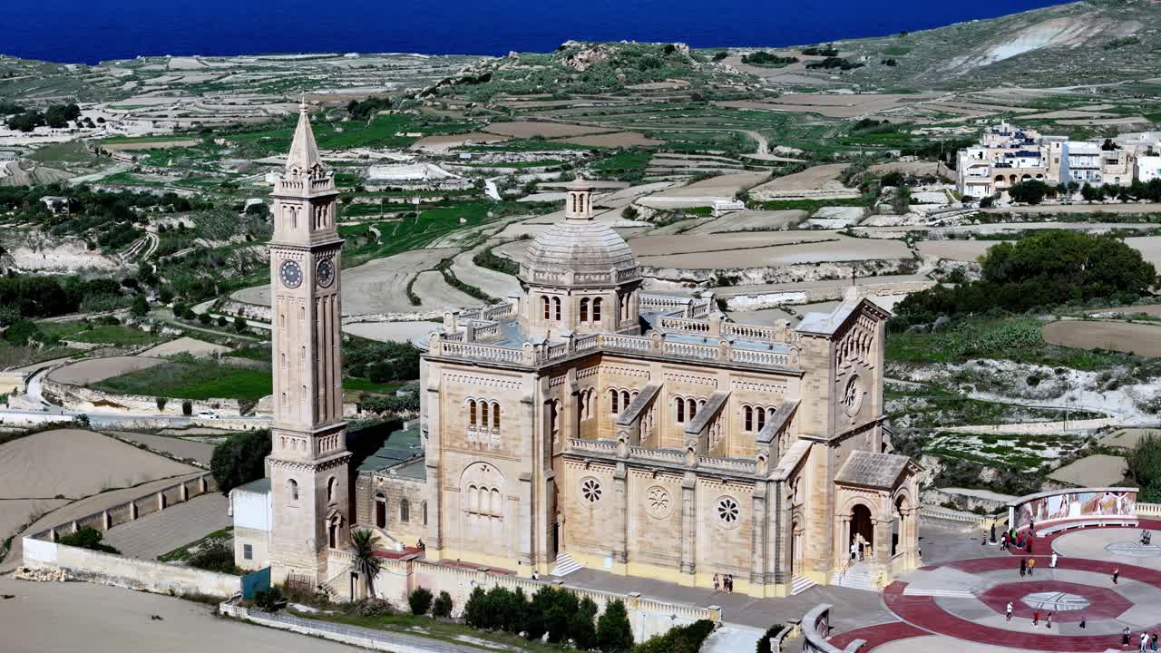 Witness the divine neo-Romanesque Ta' Pinu Basilica in Gozo Malta under a clear summer sky where golden limestone architecture meets scenic green fields in a breathtaking high-angle aerial view