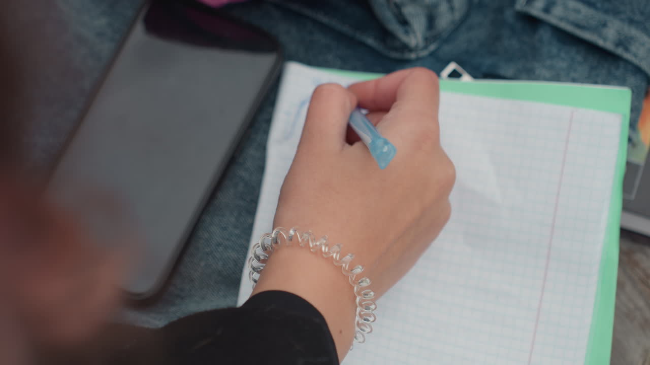 Student writing notes on grid paper, blue pen and bracelet visible beside phone and denim jacket on wooden bench, calm outdoor study atmosphere showing planner and quick sketches frames suggest