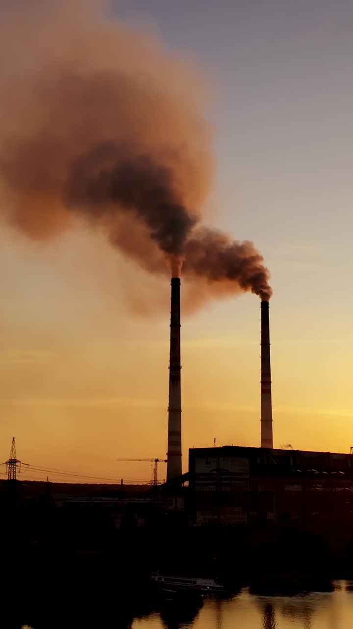 Dark power plant at the bank of the evening river. Industrial pipes of manufacturing with dark smoke at sunset. Aerial view. Vertical video