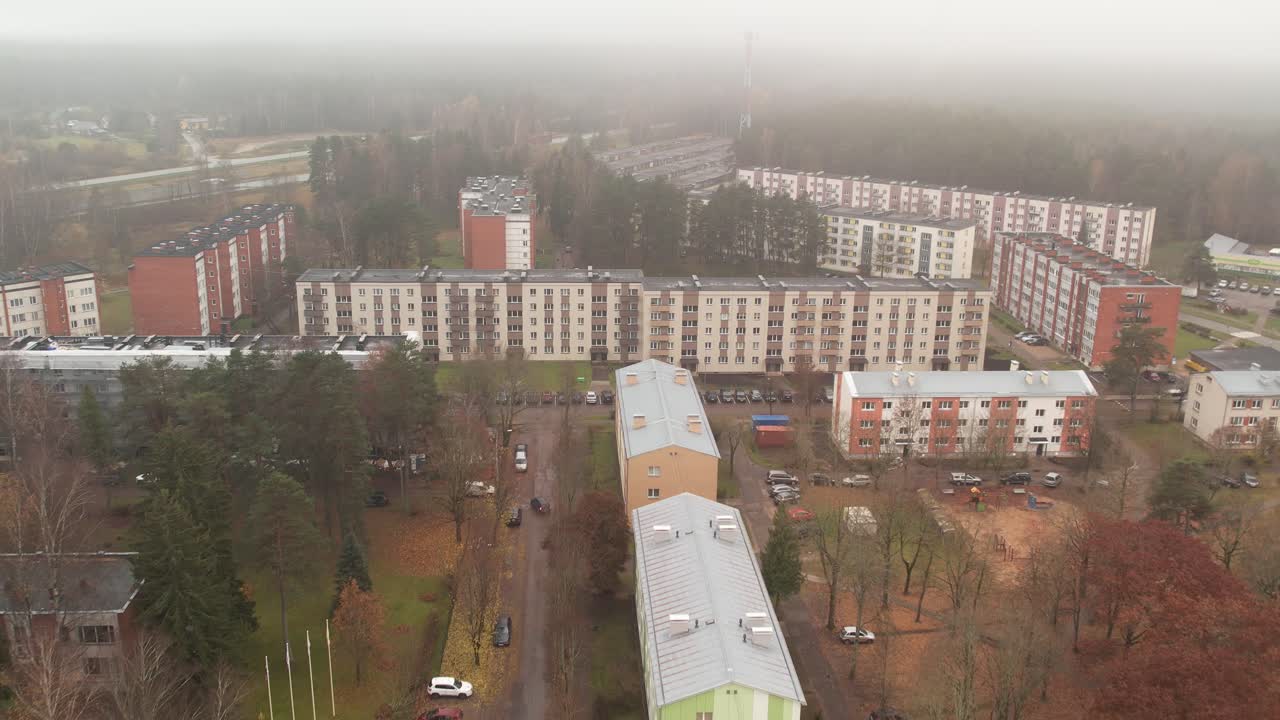 A misty aerial scene of a small town with colorful rooftops, residential buildings, and surrounding trees partially obscured by thick fog