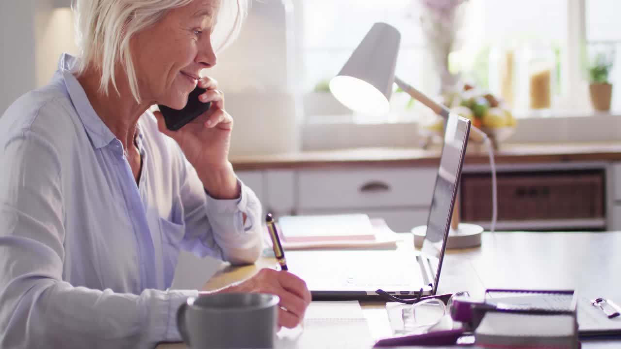 Happy senior caucasian woman sitting at table in kitchen, using laptop and talking on smartphone