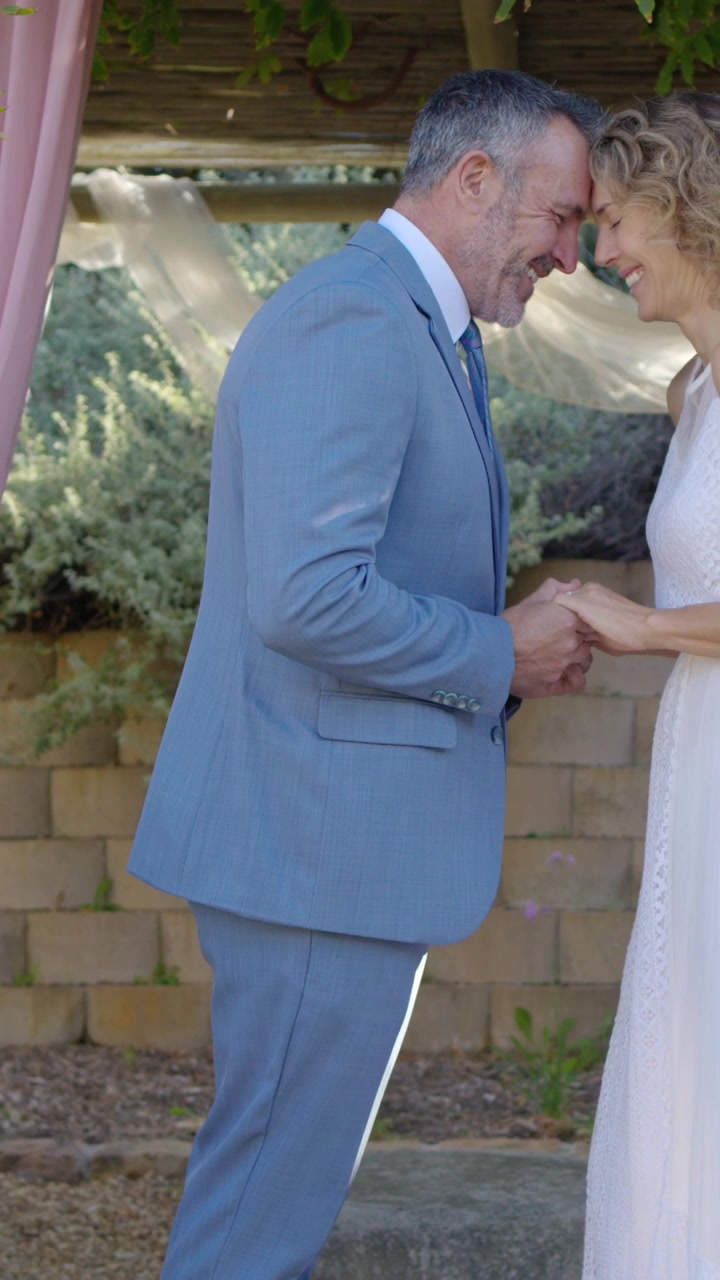 Video vertical: Novio con traje azul y novia con vestido blanco pronunciando los votos, tomados de la mano bajo una pérgola.