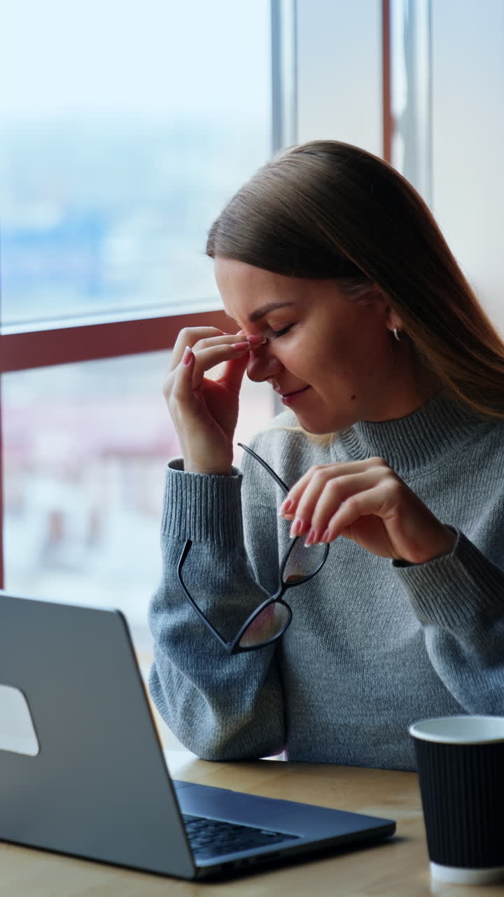 Long haired lady is tired from work at laptop. Woman took off her glasses and rubs her nose bridge. Office employee takes a break and looks at window. Vertical video