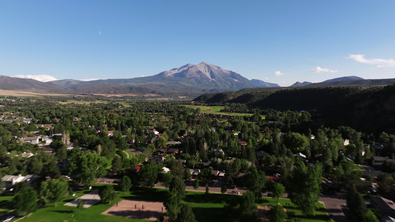 Drone fast panoramic dolly over golf course and homes of Carbondale Colorado tree lined blocks opening toward distant Mount Sopris under bright skies