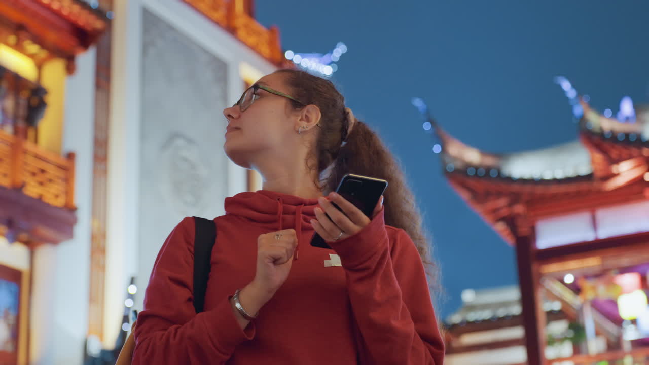 Giovane donna con felpa rossa che invia messaggi sotto le luci ornate di un tempio. Un primo piano spontaneo della viaggiatrice che scorre il telefono, illuminata dal caldo bagliore al neon delle lanterne. Lo zaino è visibile, immersa in un'atmosfera vivace di mercato notturno, con un morbido bokeh.