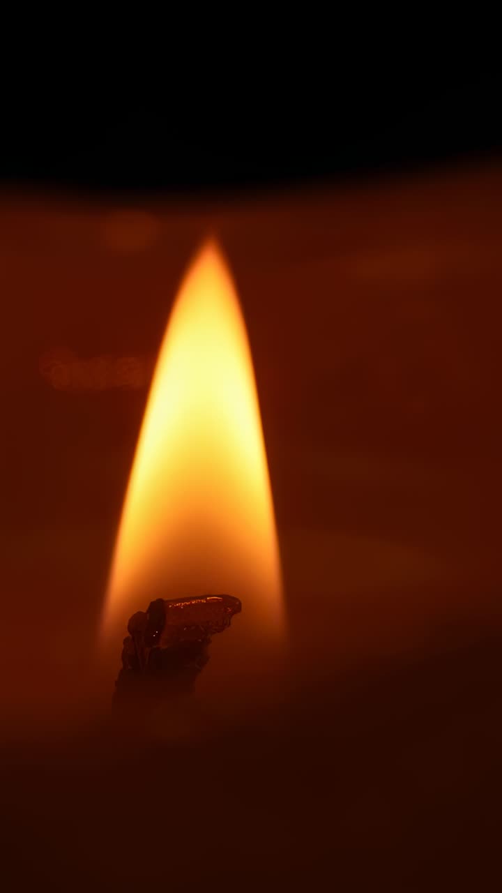 Vertical video: Burning candle flame drawing melted wax around burned wick inside dark room