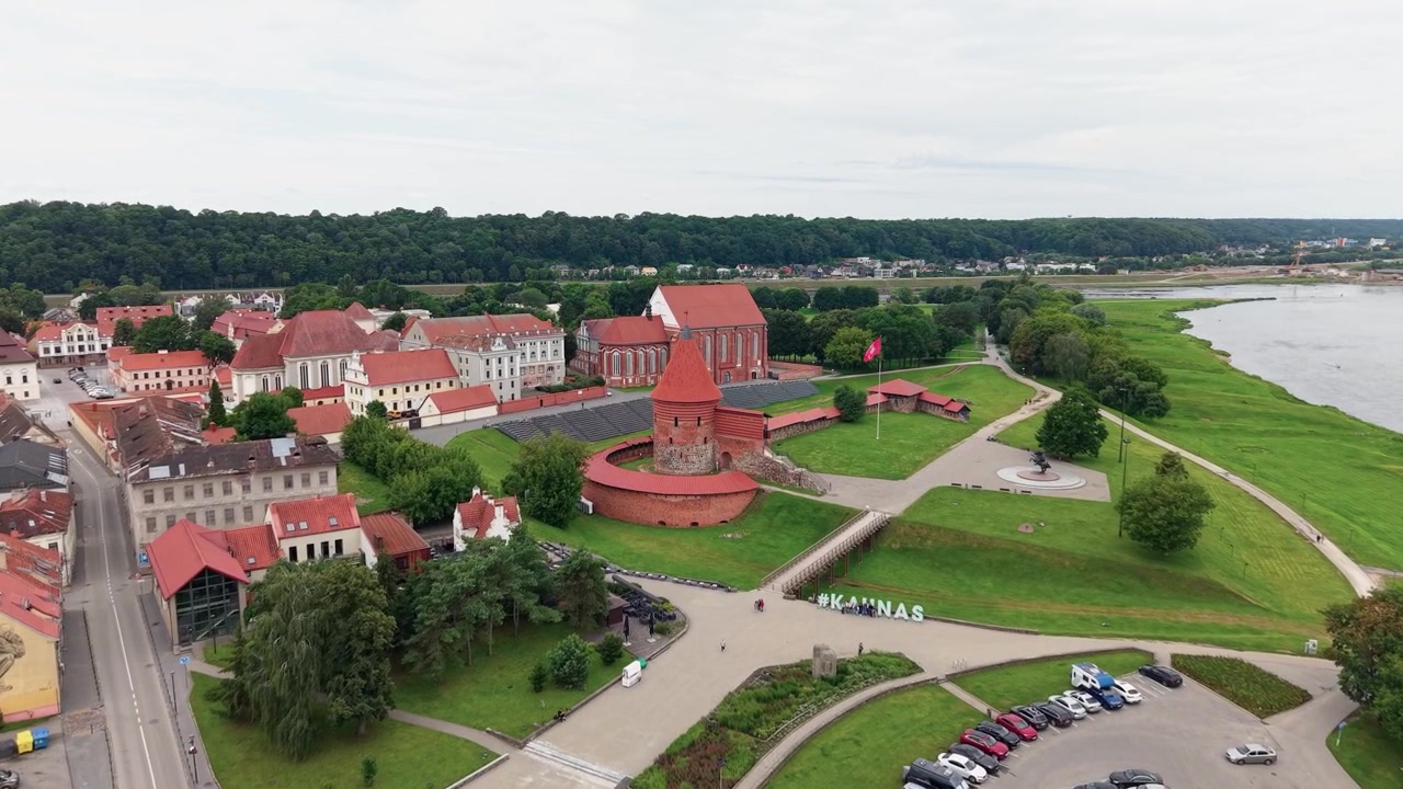 Deslumbrante filmagem aérea da Cidade Velha de Kaunas e do icônico Castelo de Kaunas de tijolos vermelhos à beira do rio, capturada em um dia nublado. Ampla tomada de drone destaca a arquitetura histórica e a vegetação exuberante.