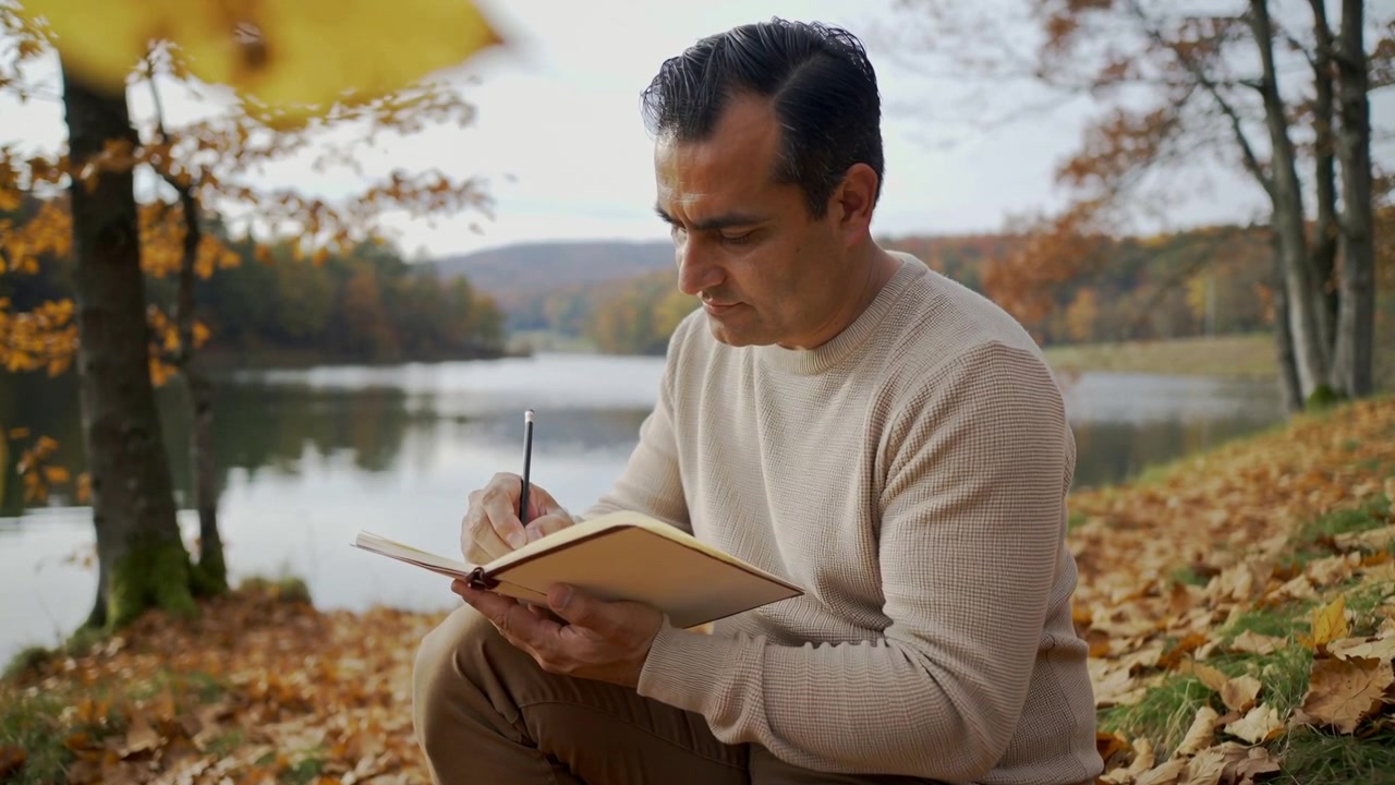 Midaldrende mand placerer notesbog på knæet, skriver dagbog ved søbredden, holder kuglepen i beige sweater.