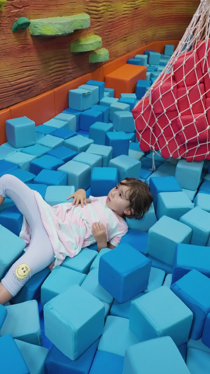 A cheerful preschool child enjoys relaxing among a large pile of varying shades of blue soft foam cubes at an indoor amusement center