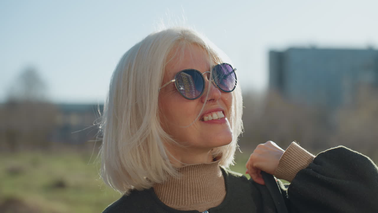 Donna bionda sorridente con capelli corti che indossa occhiali da sole e giacca scura all'aperto in un ambiente cittadino soleggiato, che si gode un momento di stile di vita con un'espressione rilassata e allegra mentre guarda verso l'alto